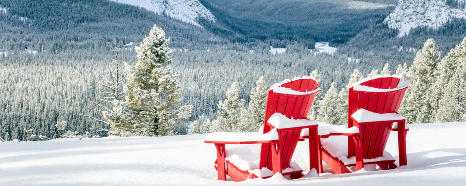 Two red Adirondack chairs covered in snow in front of a forested valley.