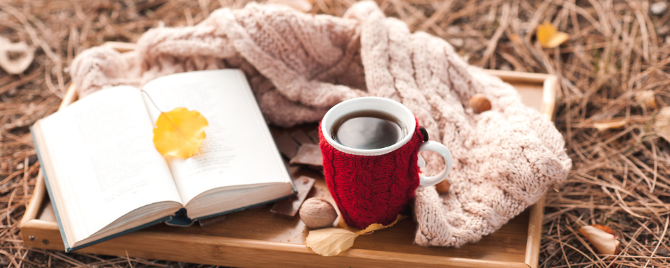 A tray holding a book, scarf, and cup of coffee sits on a bed of dry leaves.