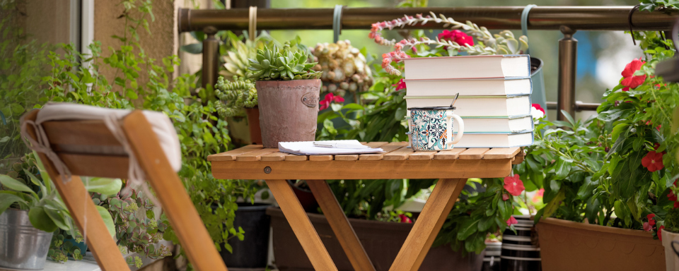 Des livres et une tasse de café se trouvent sur une petite table en bois sur une terrace, le tout entourré de plantes en pot.