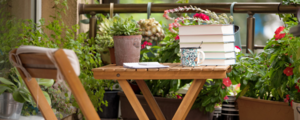 Books and a mug of coffee rest on a small wooden table on a balcony surrounded by potted plants.