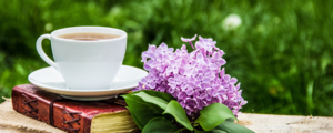 A cup of tea rests on a book beside a sprig of lilac.